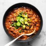 A spoon digging into a bowl of beef chili topped with sliced jalapenos.