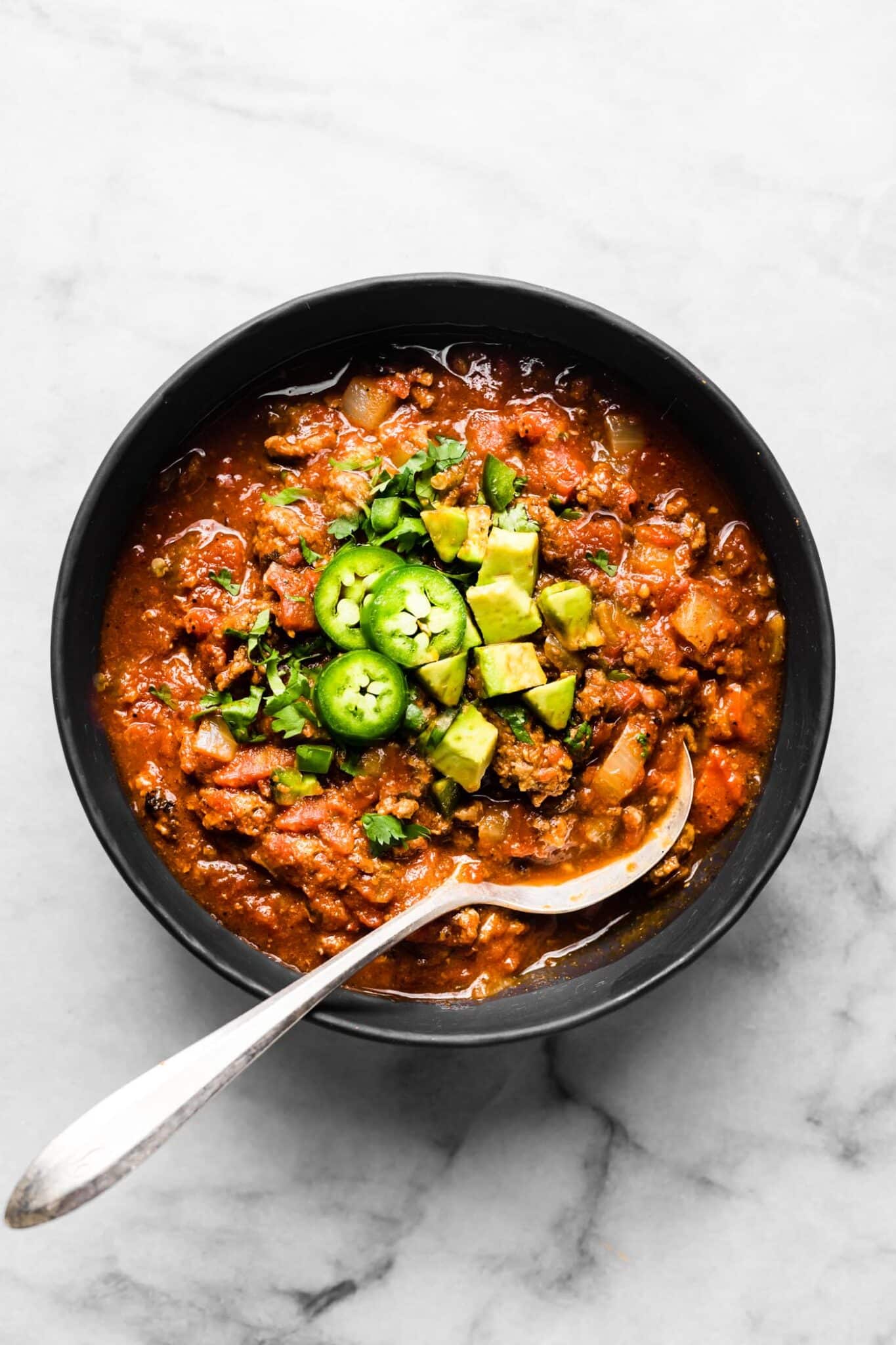 A spoon digging into a bowl of beef chili topped with sliced jalapenos.