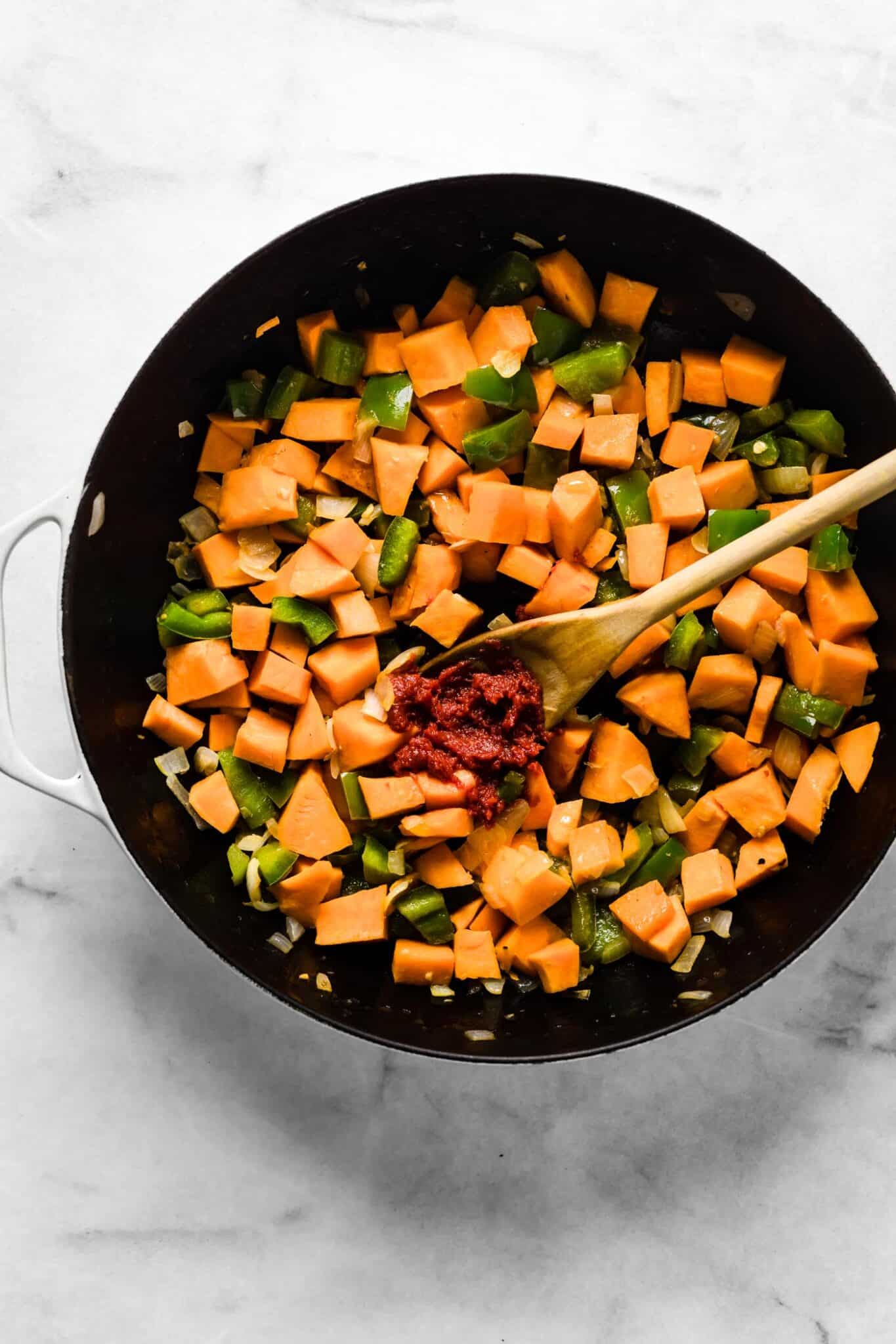 Tomato paste on a wooden spoon on top of softened vegetables in a pan.