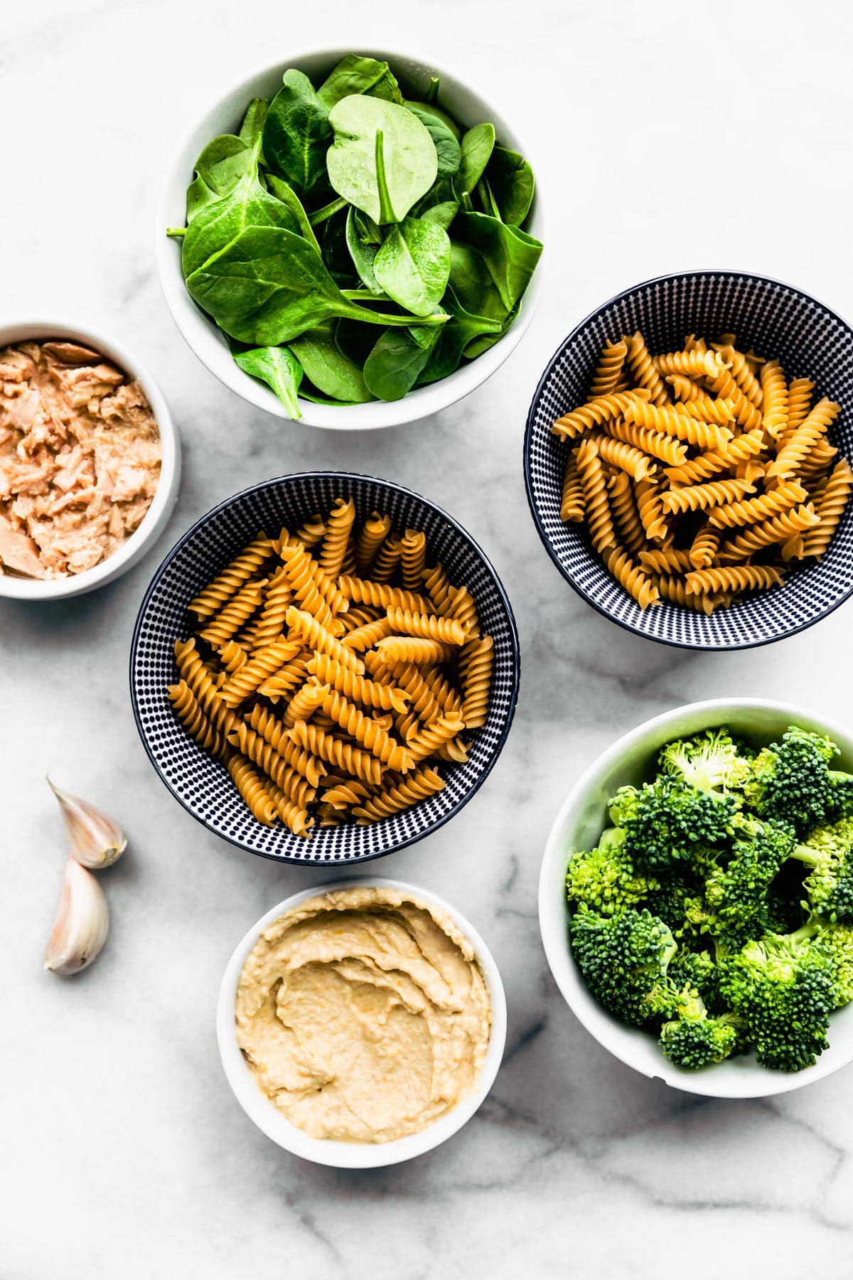Overhead shot of The Best Tuna Noodle Casserole ingredients: tuna, hummus, spinach, broccoli, and pasta in separate bowls.