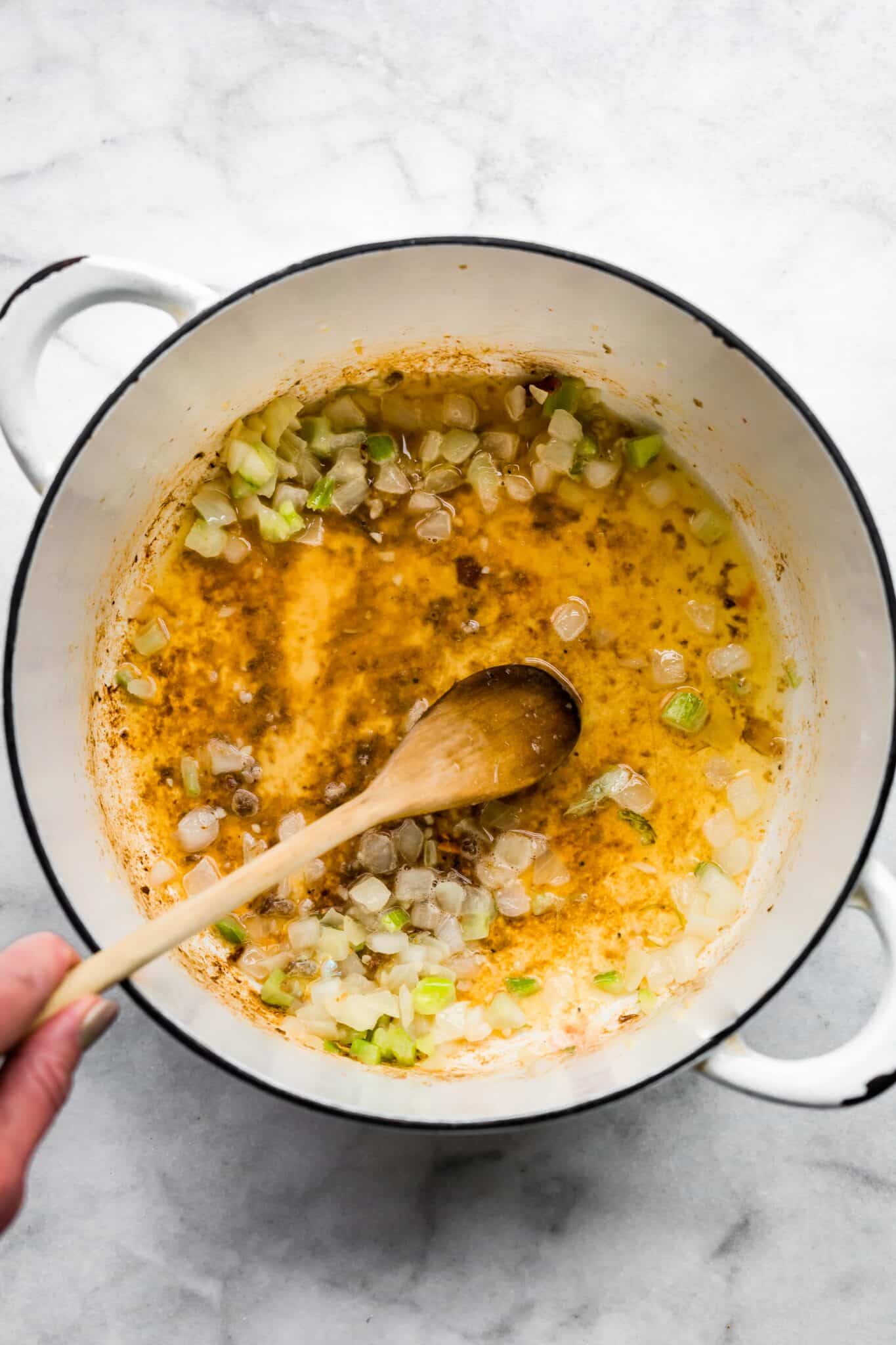 Overhead photo of a wooden spoon stirring sauteed onions and celery in a white pot.