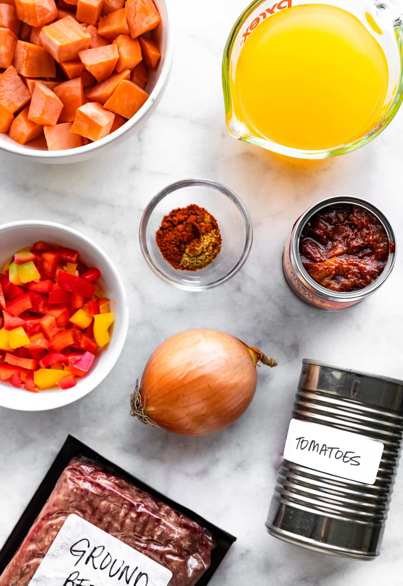 Canned tomatoes, ground beef, diced sweet potatoes, canned chipotle peppers on a white marble countertop.