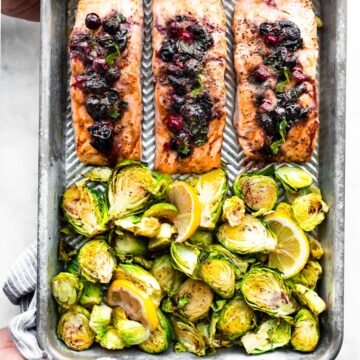 A sheet pan with three salmon filets topped with a blueberry mixture and Brussels sprouts.