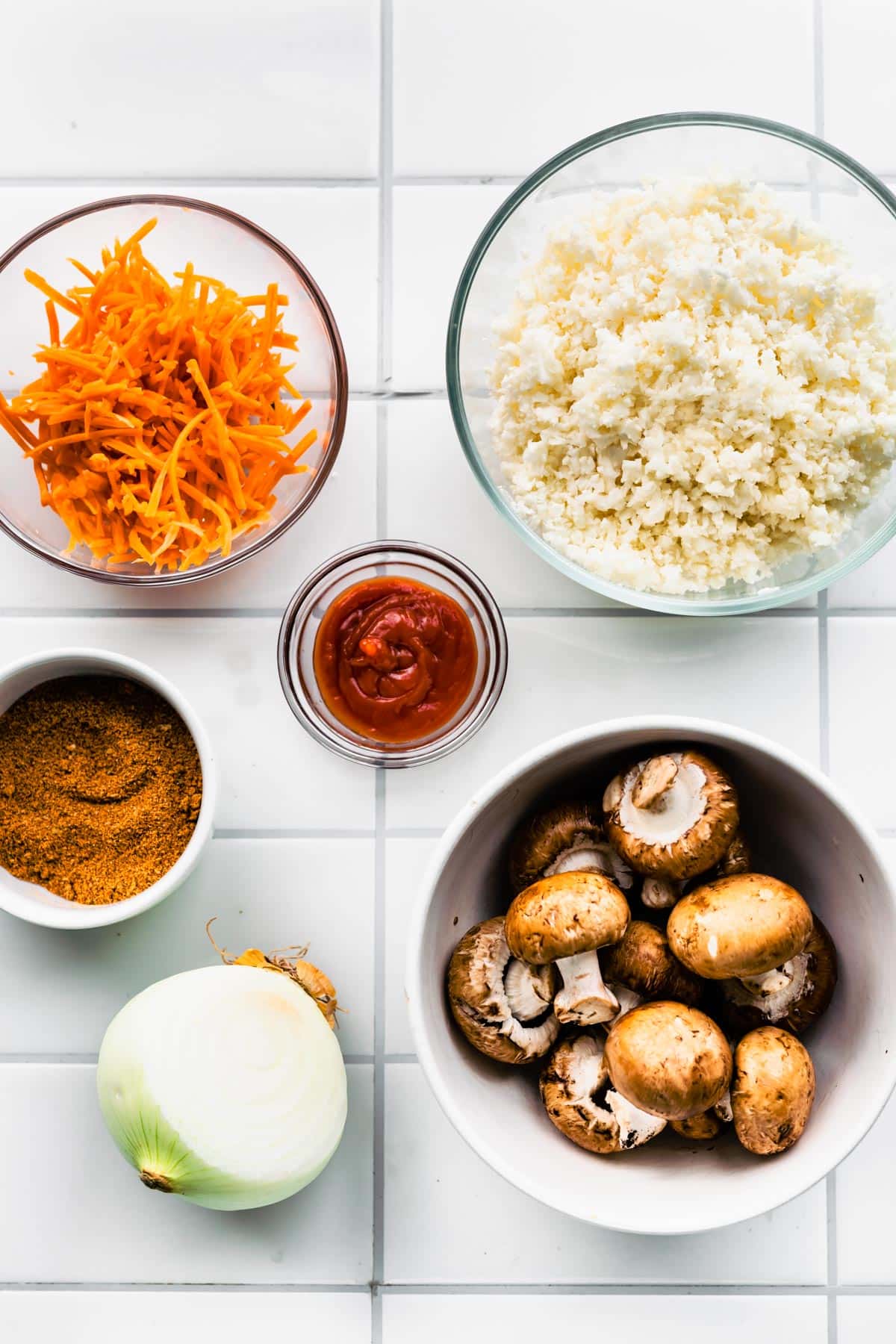 Separate bowls of mushrooms, riced cauliflower, shredded carrots and tomato paste on white countertop.