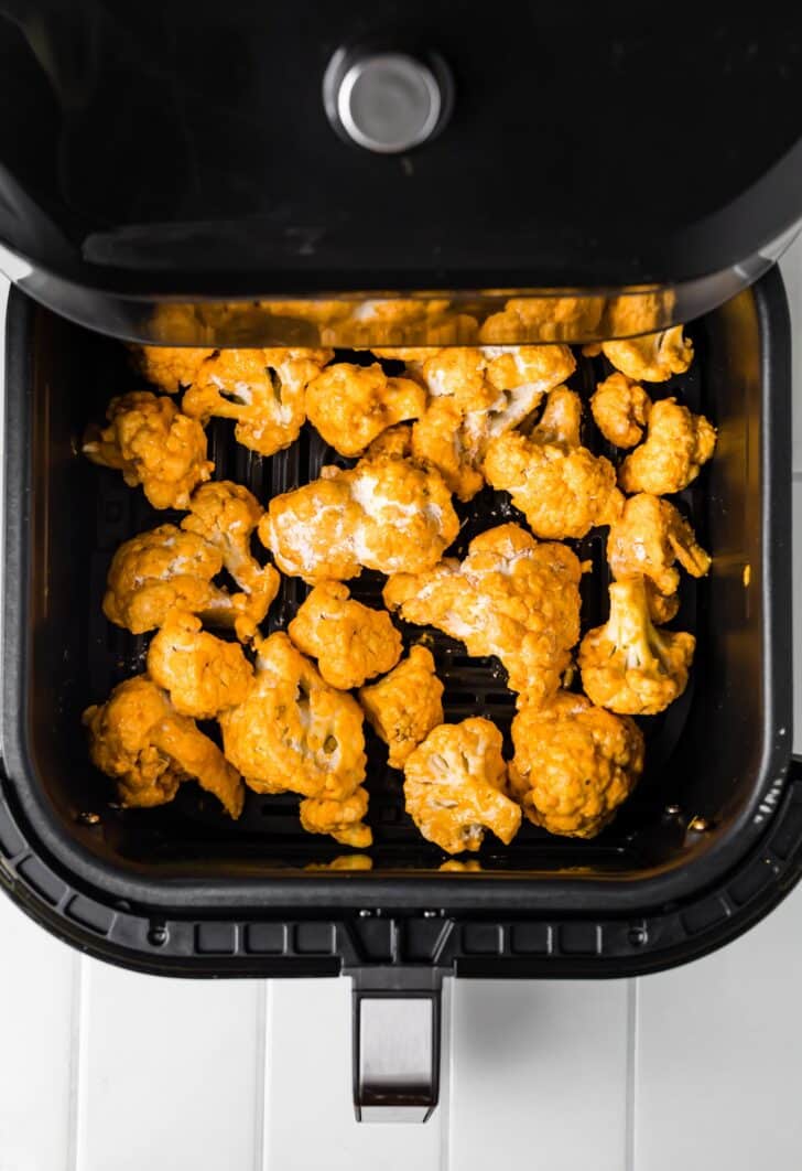 Partially fried buffalo sauce coated cauliflower in the basket of an air fryer.