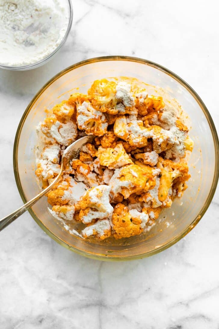 Buffalo sauce coated cauliflower in a bowl with a flour mixture on top.