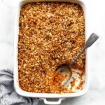 Sweet potato casserole in a white baking dish, topped with a golden nutty crumble. A section is scooped out, with the spoon resting in the empty spot. A blue-and-white striped linen sits softly scrunched along the side.