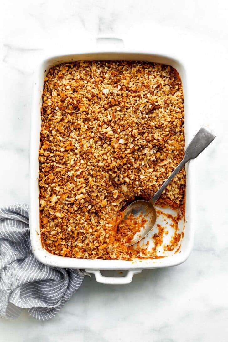 Sweet potato casserole in a white baking dish, topped with a golden nutty crumble. A section is scooped out, with the spoon resting in the empty spot. A blue-and-white striped linen sits softly scrunched along the side.